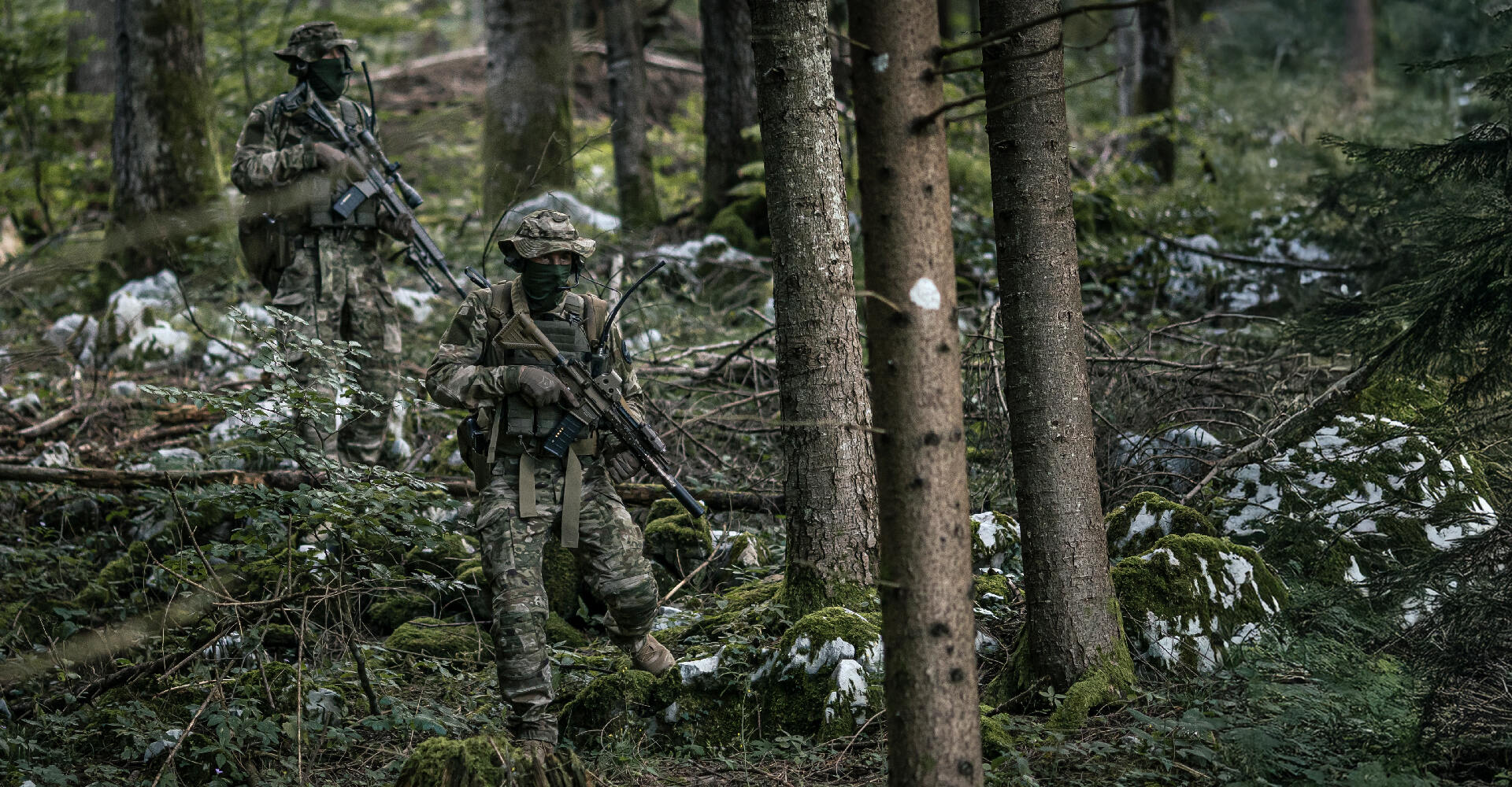 Two operators moving cautiously through the woods, blending into their surroundings with camouflage clothing.